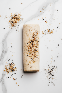This image shows dough shaped into a log and rolled in extra seeds on a lightly floured white marble surface.