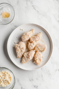 This image shows dredged chicken pieces coated and ready to cook on a white plate over a clean white marble surface.