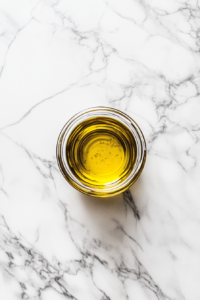This image shows a top-down view of a small clear glass bowl on a white marble countertop, containing thinly sliced pitted Kalamata olives, ready to be added to the salad