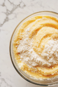 This image shows dry ingredients being gradually folded into a wet batter mixture in a large glass bowl on a white marble countertop.