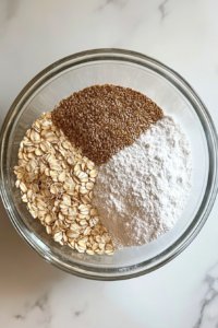This image shows a clear glass mixing bowl containing a blend of whole wheat flour, all-purpose flour, salt, oats, and assorted seeds.