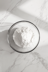 This image shows a glass bowl on a white marble surface with flour, baking soda, and salt thoroughly combined.