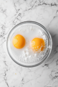 This image shows eggs and milk slightly whisked together in a glass bowl placed on a white marble surface.