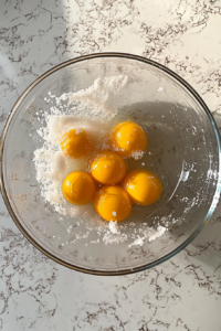 This image shows a pale, frothy mixture of egg yolks, sugar, and salt in a glass bowl on a clean white marble surface.