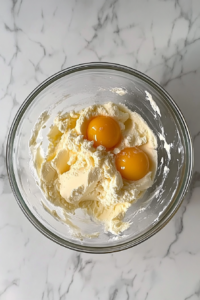 This image shows cracked eggs and vanilla extract added to a glass bowl with creamed butter and sugar, sitting on a white marble countertop.