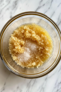 This image shows a glass mixing bowl containing separate ingredients—eggs, granulated sugar, light brown sugar, vanilla extract, and salt—on a white marble cooktop.