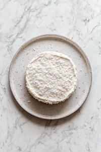 This image shows a top-down view of the first cake layer frosted and sprinkled with flaked coconut on a platter over a white marble countertop.