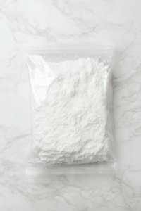 This image shows a large clear plastic bag partially filled with white all-purpose flour, lying open on a clean white marble countertop, with no other items or ingredients in the background.