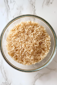 This image shows fluffed brown rice cooling in a large clear glass bowl placed on a white marble cooktop.