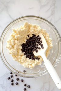 This image shows a glass bowl on a white marble countertop with cookie dough, milk, and chocolate chips being folded in using a white rubber spatula.