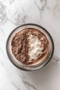 This image shows a chocolate cake batter being lightened with whipped egg whites in a glass bowl on a white marble countertop.