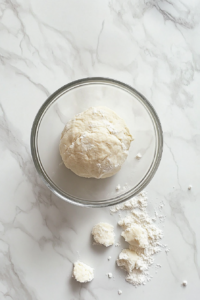 This image shows a sticky dough ball forming in a glass bowl, surrounded by small spoonfuls of flour over a white marble countertop.