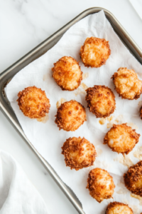 This image shows several pieces of crispy fried chicken resting on white paper towels placed over a silver baking sheet, set on a clean white marble countertop with no other items in the background.