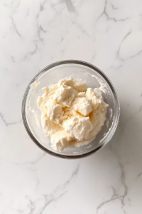 This image shows a top-down view of creamy frosting in a glass bowl made by beating sugar and cream into butter, on a white marble countertop.