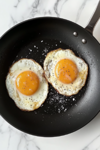 This image shows sunny-side-up eggs being fried in a black nonstick skillet on a white marble cooktop, yolks seasoned with salt and black pepper.