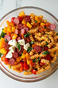 This image shows a fully tossed rotini pasta salad in a glass bowl, evenly coated with Italian dressing, resting on a white marble cooktop.