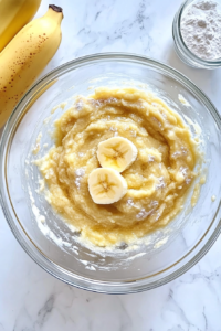 This image shows a large glass mixing bowl containing mashed bananas, flour, sugar, butter, egg, vanilla, baking soda, and baking powder, placed alone on a clean white marble countertop.