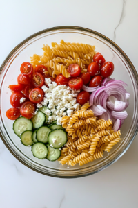 This image shows a clear glass bowl on a white marble surface containing pasta, tomatoes, cucumber, red onion, olives, and feta, ready to be mixed for Greek pasta salad.