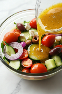 This image shows a glass bowl of Greek pasta salad with vinaigrette poured on top, about to be tossed, sitting on a white marble countertop with clean background.