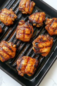 This image shows marinated chicken thighs grilling on a black cast iron pan with char marks, over a white marble countertop.