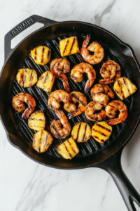 This image shows shrimp and pineapple slices grilling on a black cast iron grill pan over white marble cooktop. The shrimp are pink and seared, and the pineapple has visible grill marks.