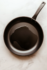 This image shows oil being heated in a large black nonstick skillet on a white marble cooktop.