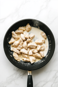 heavy-whipping-cream-pouring-into-black-skillet-with-cooked-chicken-on-white-marble