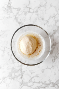 This image shows a smooth dough ball coated in oil placed inside a greased glass bowl on a white marble surface.