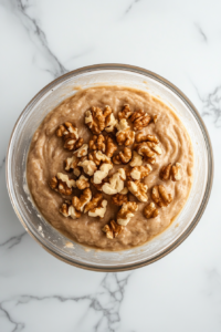 This image shows thick apple cake batter with chopped walnuts folded into it inside a clear glass bowl on a white marble countertop.