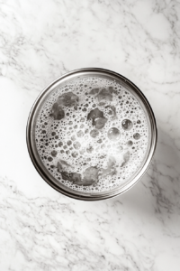 This image shows a top-down view of a large stainless steel pot filled with boiling water and salt, placed on a white marble cooktop, with no other items in the background.