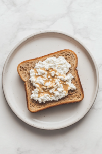 This image shows a top-down view of a slice of toasted multigrain bread on a matte white ceramic plate over a clean white marble countertop, with a thin layer of smooth peanut butter spread directly onto the toast under the layer of white low-fat cottage cheese.