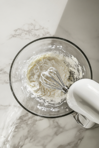 This image shows a clear glass mixing bowl with eggs and sugar being whipped to a thick texture using a stand mixer, placed on a white marble cooktop.