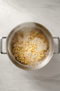 This image shows elbow macaroni boiling in salted water inside a stainless steel pot on a clean white marble cooktop, with bubbles and steam rising.