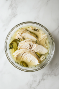 This image shows chicken slices marinating in a mixture of pickle juice and milk in a clear glass bowl on a clean white marble surface.