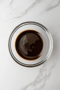 This image shows a glossy chocolate and butter mixture cooling in a glass bowl placed on a white marble countertop.