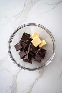 This image shows a glass ovenproof dish with semi-sweet chocolate and butter melting gently on a clean white marble cooktop.
