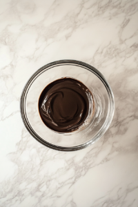 This image shows a clear glass bowl on a white marble cooktop, holding chopped German sweet chocolate and water, melted together smoothly.