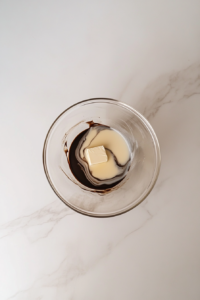 This image shows a clear glass bowl containing melted chocolate, butter, and cream for ganache preparation on a white marble cooktop.