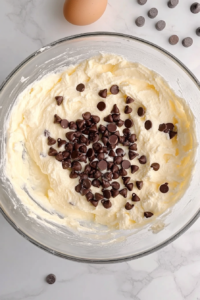 This image shows a top-down view of mini chocolate chips evenly stirred into pound cake batter in a clear glass bowl on a marble countertop.