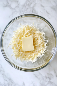 This image shows a glass mixing bowl filled with butter, minced garlic, Italian dressing, and mozzarella cheese on a white marble cooktop.