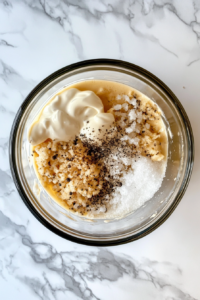 This image shows a clear glass bowl containing a creamy mixture of Hellmann’s mayonnaise, vinegar, salt, sugar, and pepper on a clean white marble surface.