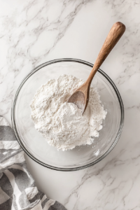 This image shows a top-down view of a glass bowl on a white marble cooktop where flour, baking powder, and salt are being mixed into the peanut butter batter.