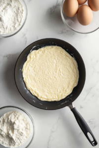This image shows a black nonstick skillet with melting butter and freshly poured crepe batter, next to a glass bowl of batter on a white marble cooktop.