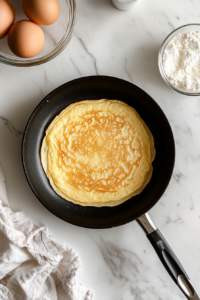This image shows crepe batter evenly spread in a black nonstick skillet after tilting, with a glass bowl of remaining batter on a white marble cooktop.