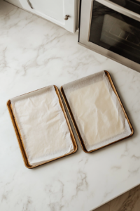 This image shows a top-down view of two parchment-lined baking trays on a white marble countertop with the oven preheating in the background.