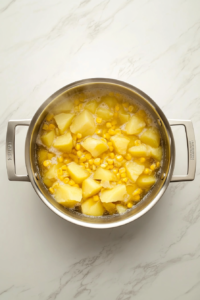 This image shows the corn and potato soup simmering in a stainless steel pot on a clean white marble countertop.