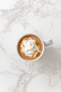 This image shows a white ceramic mug filled with freshly poured French hot chocolate, topped with whipped cream, placed on a white marble cooktop.