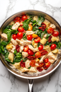 This image shows a creamy mixture of milk, flour, and Parmesan cheese poured into a stainless steel skillet over chicken, sun-dried tomatoes, grape tomatoes, artichokes, and baby spinach on a clean white marble cooktop