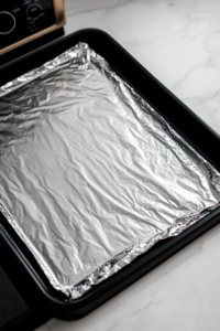 This image shows a black baking sheet lined with foil placed on a white marble countertop, with the oven preheating.