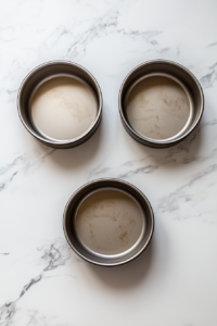 This image shows a top-down view of an oven preheating to 350°F and three greased, floured 8-inch round cake pans on a white marble countertop.